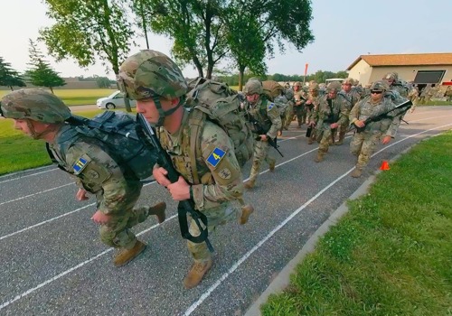 A group of Army National Guard soldiers in full gear participate in a training exercise, running on a track with backpacks and rifles.