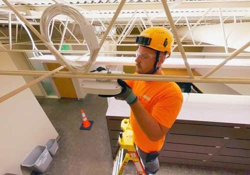 A technician wearing an orange safety shirt, helmet, and gloves installs network equipment on a ceiling while standing on a ladder in an office setting.