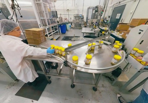 A worker in a sunflower butter facility wearing a hairnet and lab coat monitors jars with yellow lids moving along a conveyor system.